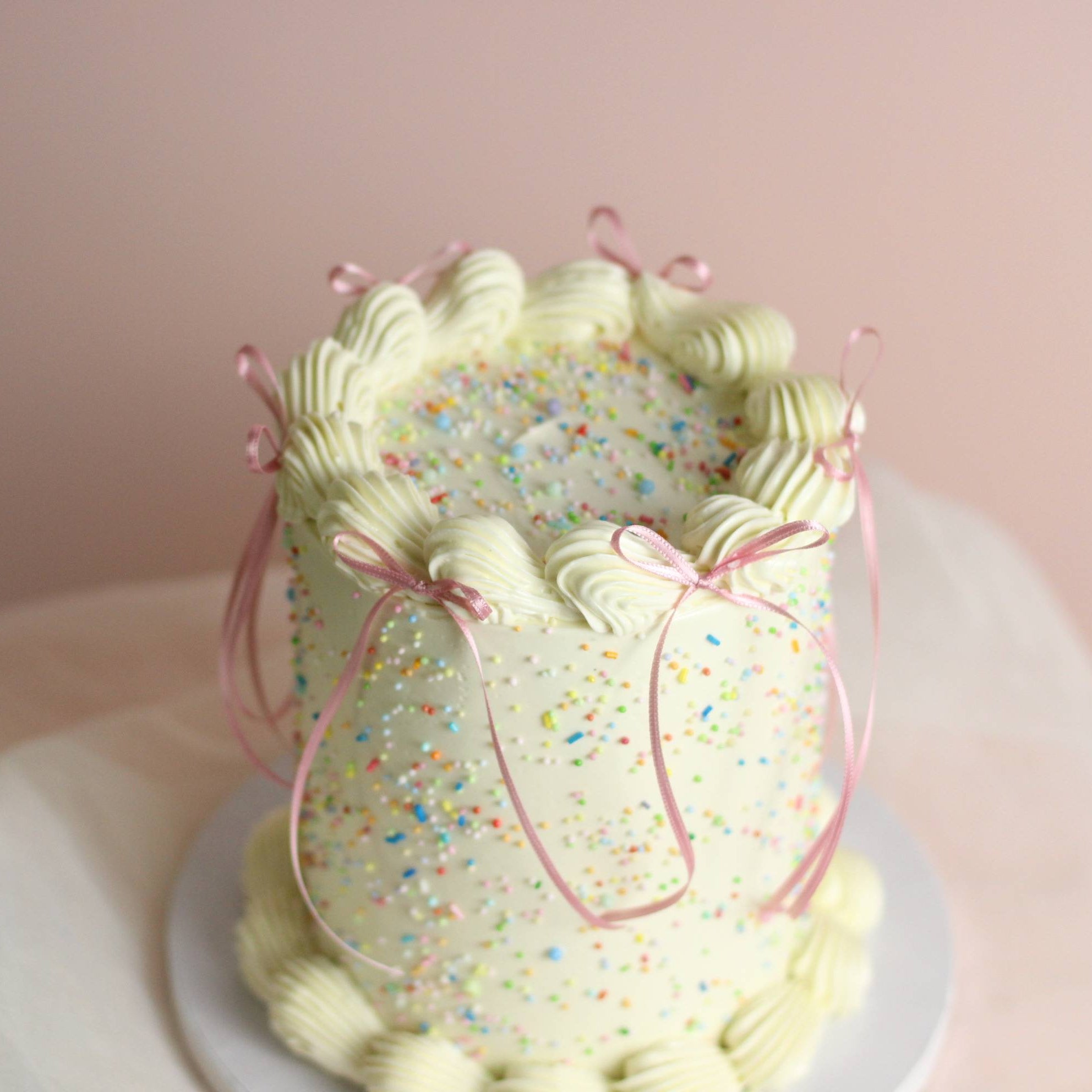 Customizable buttercream birthday cake with vintage piping, rainbow sprinkles, and ribbon bow details. Choose your preferred cake color and frill color, with an optional pastel candle add-on for an extra festive touch. Perfect for birthdays, celebrations, and special occasions in Berlin.