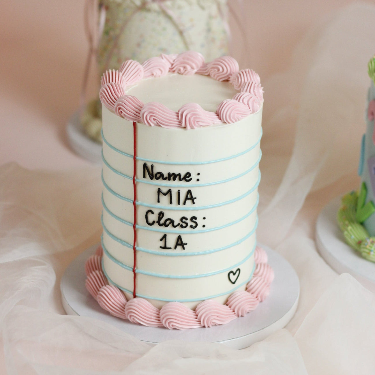 Mark their school start with this custom notebook cake. Designed with classic blue notebook lines, a red margin, and customizable text for your child’s name and class, all framed by soft vintage piping. The sweetest way to kick off the new school year!