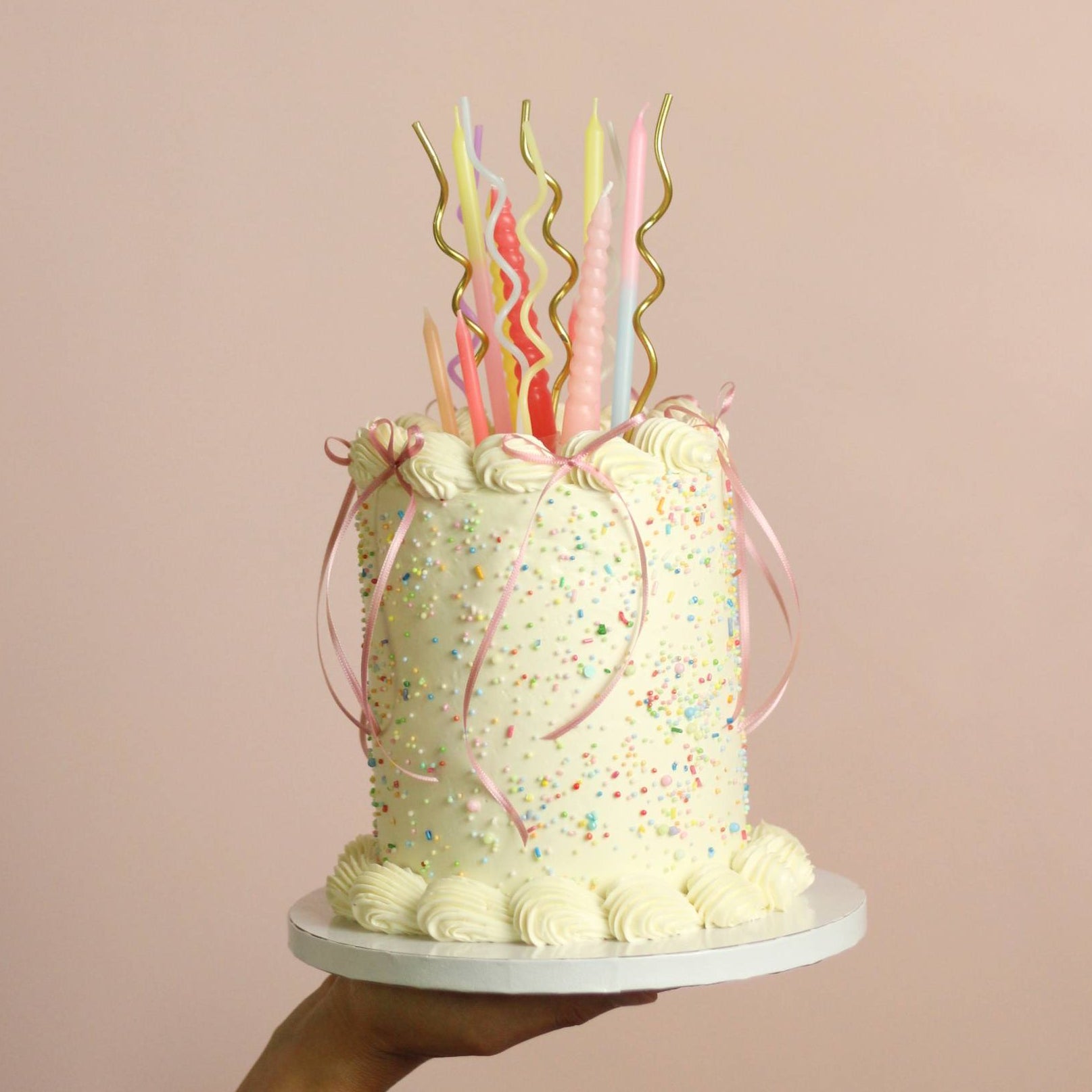 Customizable buttercream birthday cake with vintage piping, rainbow sprinkles, and ribbon bow details. Choose your preferred cake color and frill color, with an optional pastel candle add-on for an extra festive touch. Perfect for birthdays, celebrations, and special occasions in Berlin.