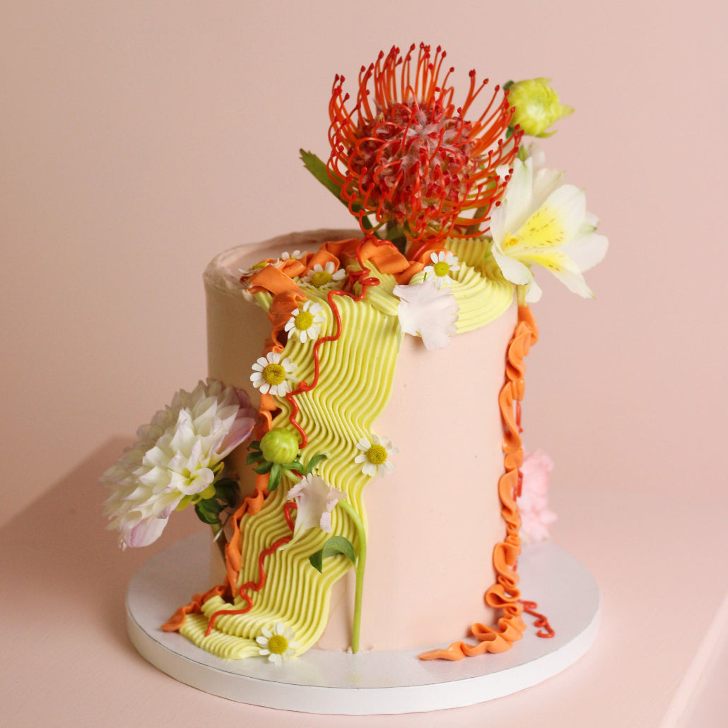 A decorated cake with a pale blush base, a yellow-green ribbed draping ribbon, orange ruffled trim, and edible flowers including a red protea-style bloom, photographed on a soft pink studio background.