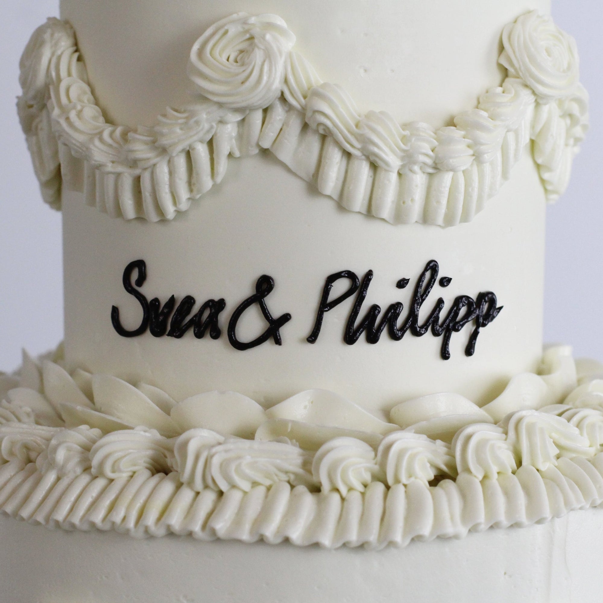 Close-up of a vintage white wedding cake with intricate buttercream frills and a custom handwritten piped message in black.